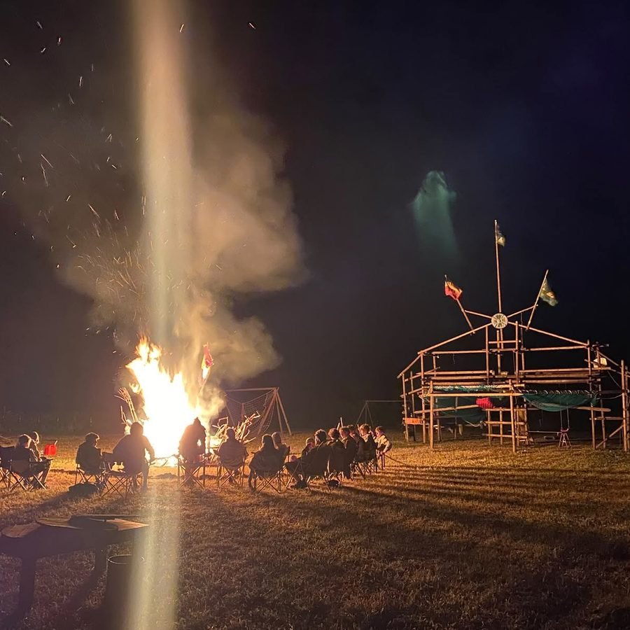 Kampvuur in de avond met scouts rondom het vuur en een kampbouw op de achtergrond